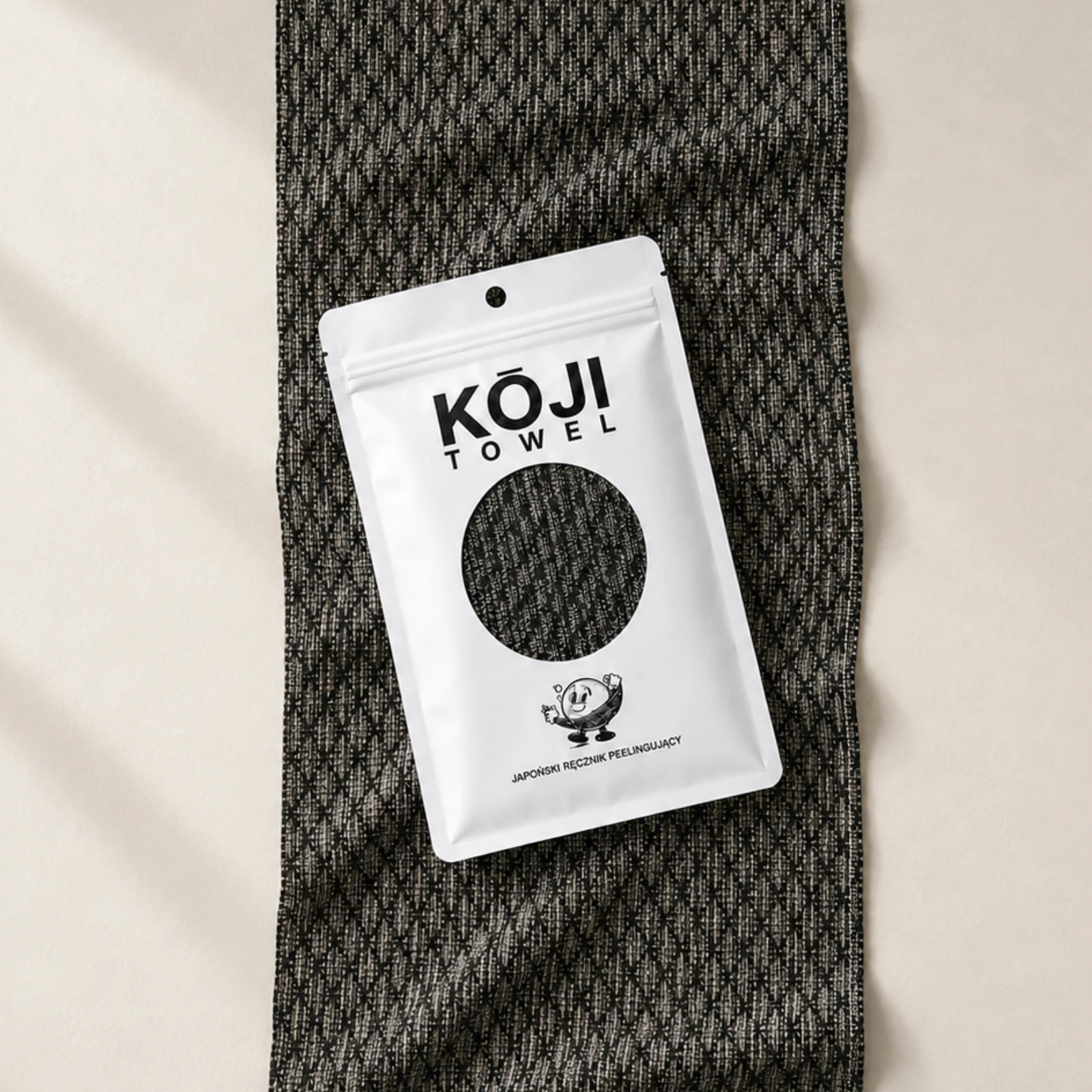 Białe opakowanie Kōji Towel ułożone na długim czarnym ręczniku